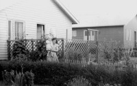 Old black-and-white photo. Two wooden houses, a lady with a child in her arms in front of one.
