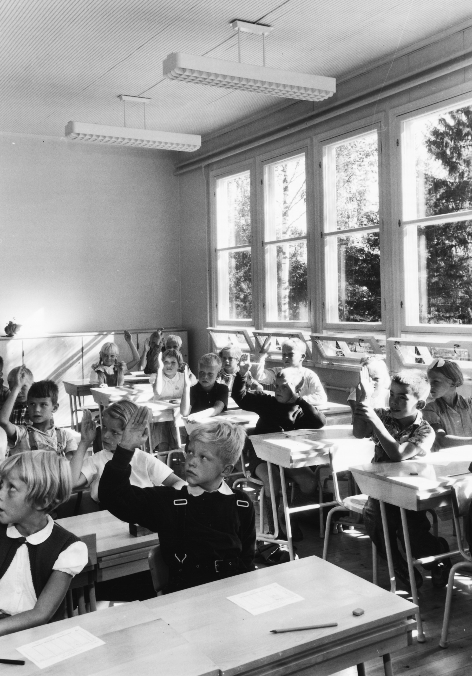 Black and white photo of Puutalo school classroom, where a class is in session. The pupils are raising their hands.