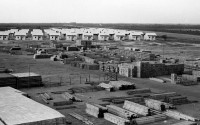 Old black-and-white photo. In the background an army of small wooden houses, in the front stacks of wooden elements.