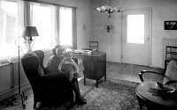 Old black-and-white photo. A living-room, windows behind an armchair with a lady and a child, two decorative lamps, wooden lacquered desk, oriental carpet.