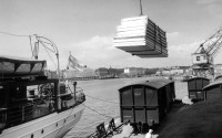 Old black-and-white photo, wooden elements being lifted from a train to a ship. Harbour crane in the background.